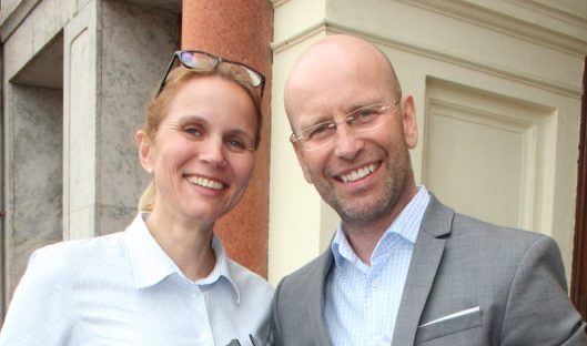 Kristina Klintö och Magnus Becker står utanför en byggnad och ler. Kristina har på sig vit skjorta samt glasögon uppskjutna på huvudet. Magnus har på sig blåvit skjorta och grå kavaj samt glasögon.